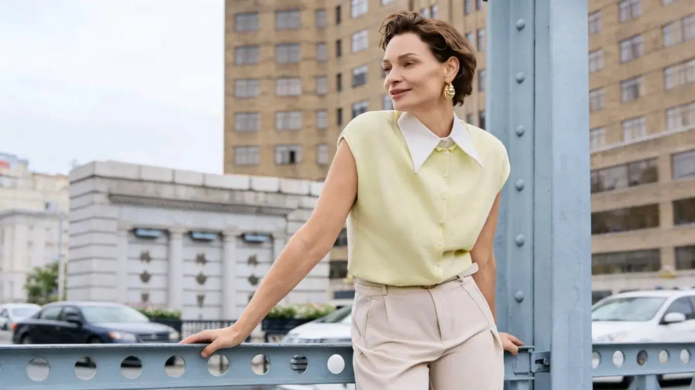 Full-body look of a lady in a light yellow sleeveless knitted top with a white fake collar, beige high-waisted wide-leg pants, and gold earrings against an urban bridge backdrop