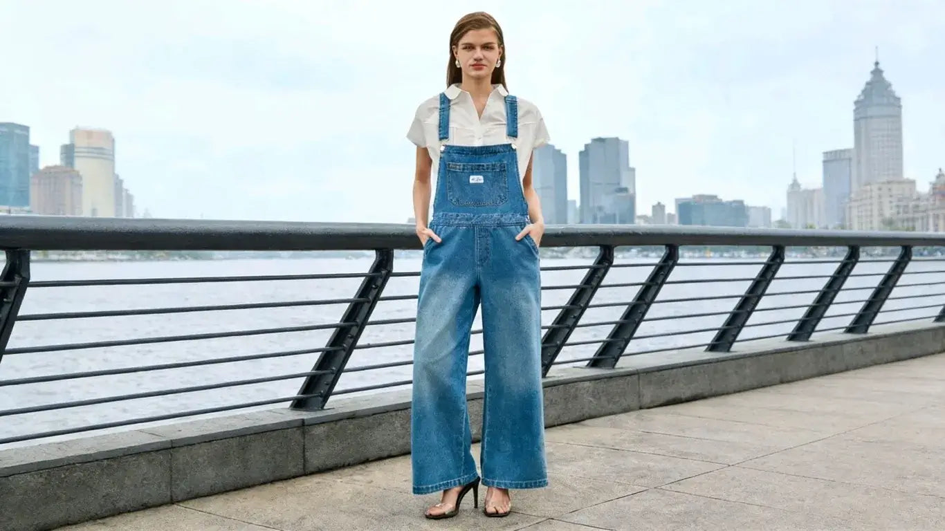  A woman wearing denim overalls stands by the riverside with city skyline in the background