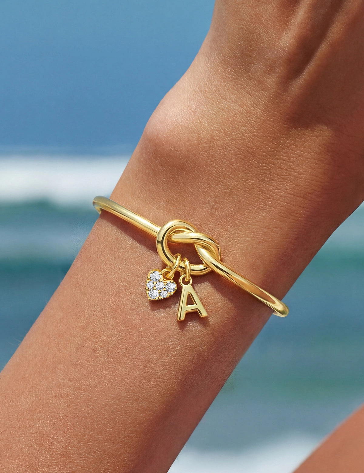 Cicy Bell Women's 18k Gold Plated Initial Bangle featuring a symbolic love knot design and a sparkling CZ heart charm. This personalized open-cuff bracelet offers an adjustable $60\text{mm}$ fit for bridesmaids or best friends.