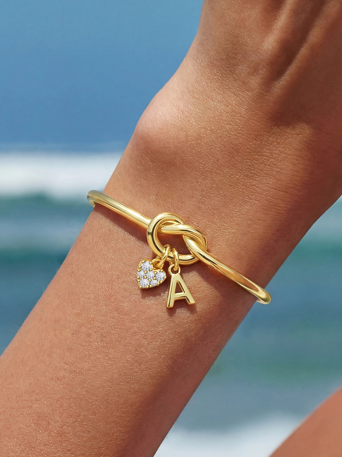 Cicy Bell Women's 18k Gold Plated Initial Bangle featuring a symbolic love knot design and a sparkling CZ heart charm. This personalized open-cuff bracelet offers an adjustable $60\text{mm}$ fit for bridesmaids or best friends.