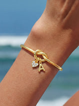 Cicy Bell Women's 18k Gold Plated Initial Bangle featuring a symbolic love knot design and a sparkling CZ heart charm. This personalized open-cuff bracelet offers an adjustable $60\text{mm}$ fit for bridesmaids or best friends.
