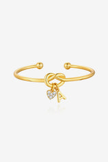 Cicy Bell Women's 18k Gold Plated Initial Bangle featuring a symbolic love knot design and a sparkling CZ heart charm. This personalized open-cuff bracelet offers an adjustable $60\text{mm}$ fit for bridesmaids or best friends.